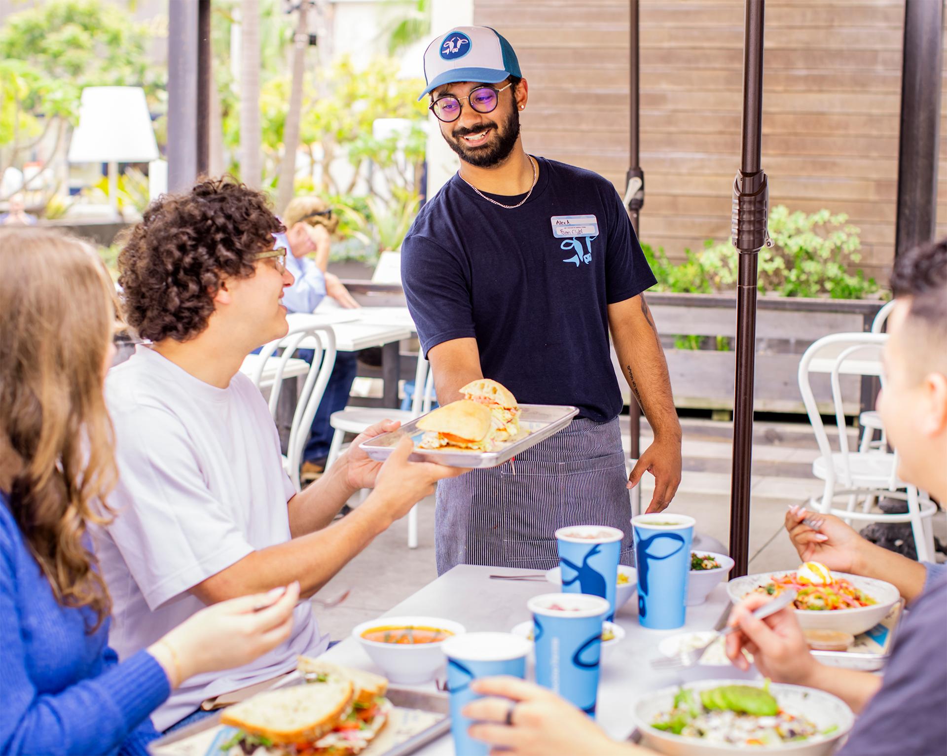 Server handing a sandwich to a smiling diner at an outdoor restaurant table, surrounded by people eating, with blue cups and plates of food.